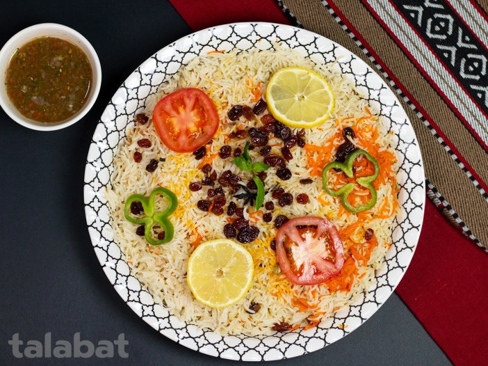 A traditional Sitra-style rice dish topped with raisins, lemon slices, tomato, chili, and carrot, served with green chutney on the side.