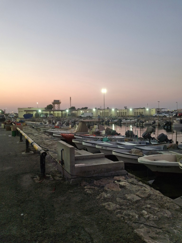 A marina at dusk with several boats docked along a stone walkway, illuminated by lights. The sunset creates a colorful sky in the background.