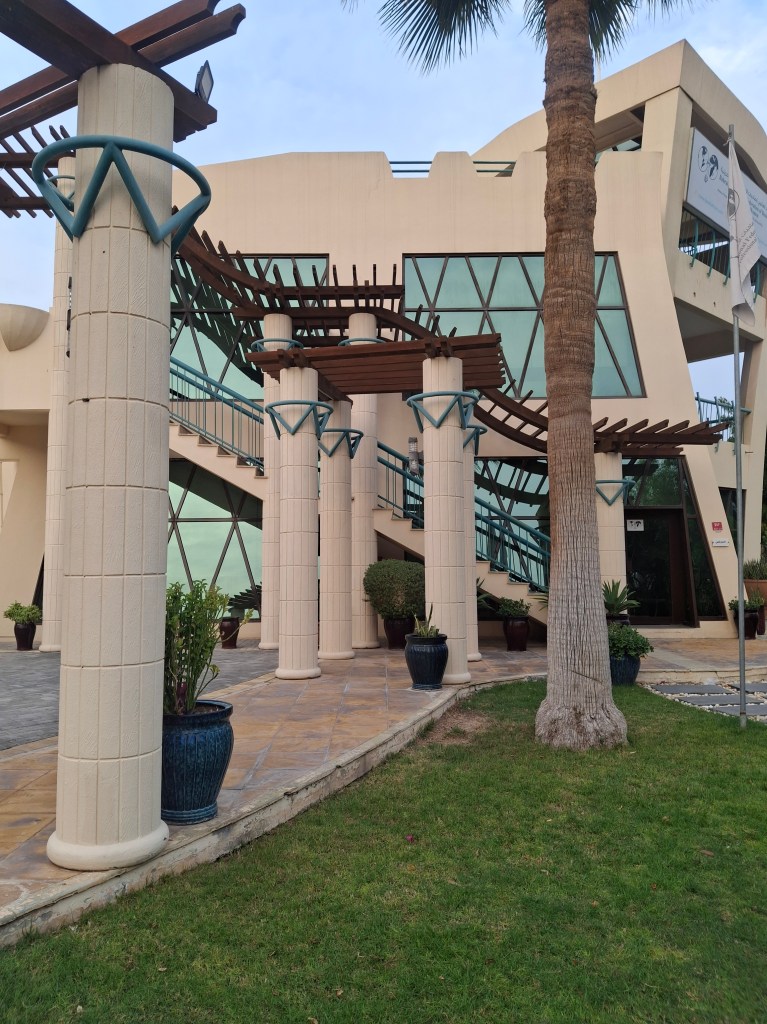 Exterior view of a modern building with unique architectural features, columns, and greenery in front, located in Bahrain.