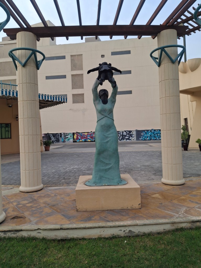 A bronze statue of a woman raising a child above her head, situated in an outdoor area with a mix of modern architecture and vibrant graffiti in the background.