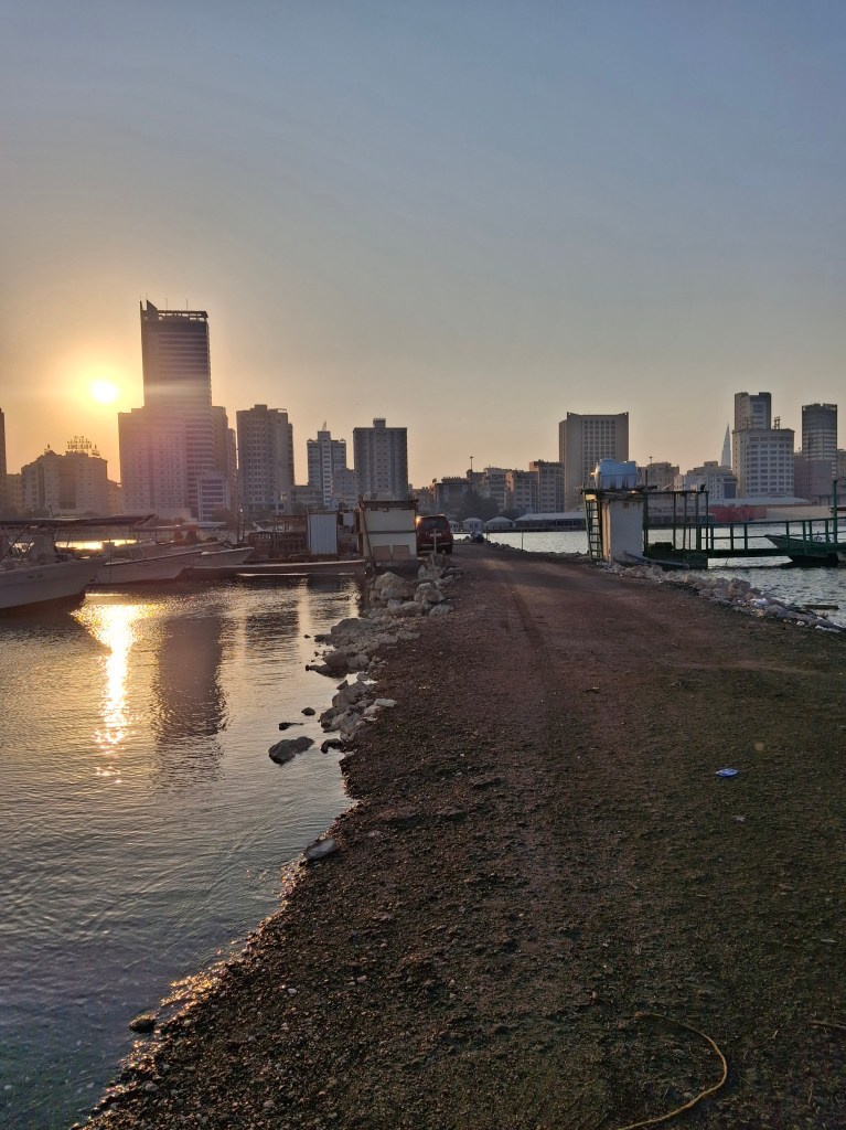 A waterfront view of Bahrain Marina during sunset, with a skyline of modern buildings illuminated by the golden sunlight.