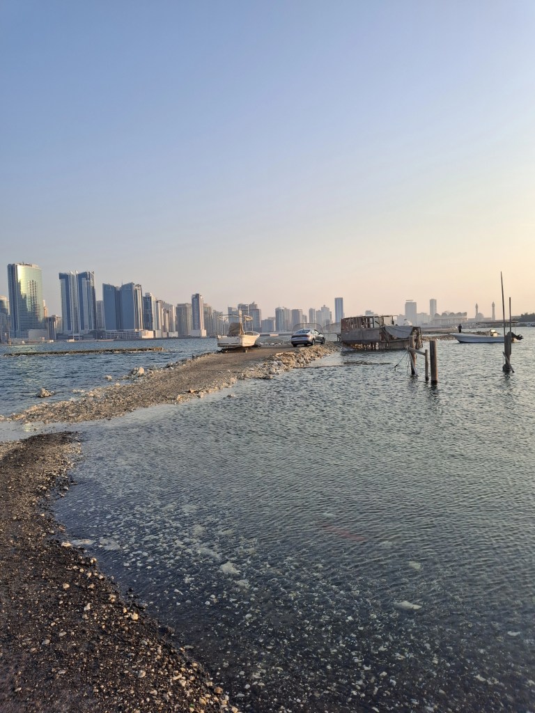 View of Bahrain's coastline featuring modern skyscrapers, a rocky shoreline, and boats anchored in shallow waters at sunset.