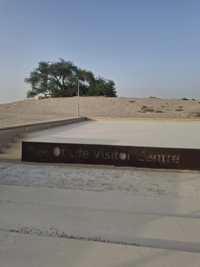 Tree of Life Visitor Centre sign in the foreground with the iconic Tree of Life on a sandy hill behind it, located near Sitra, Bahrain under a clear blue sky.