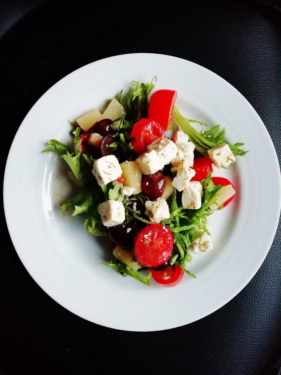 A colorful salad featuring mixed greens, cherry tomatoes, grape halves, red bell pepper, feta cheese, and chunks of pineapple, served on a white plate.