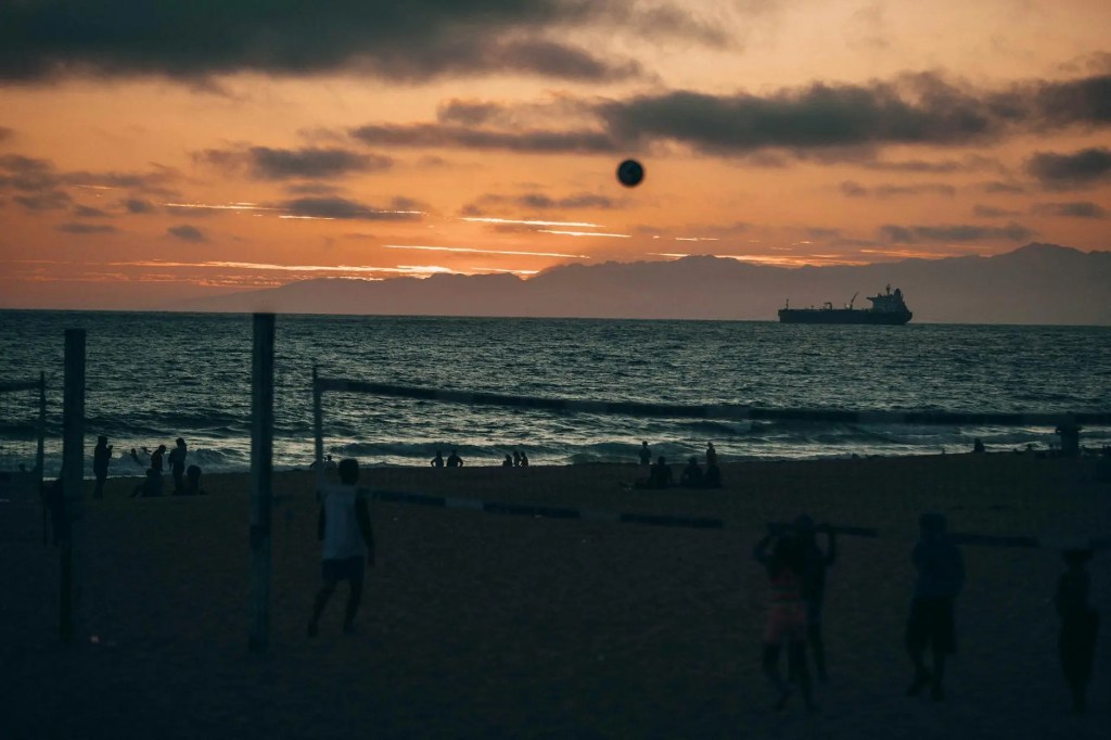 At sunset on a beach, people play volleyball and enjoy the serene ocean view with a ship sailing in the distance.