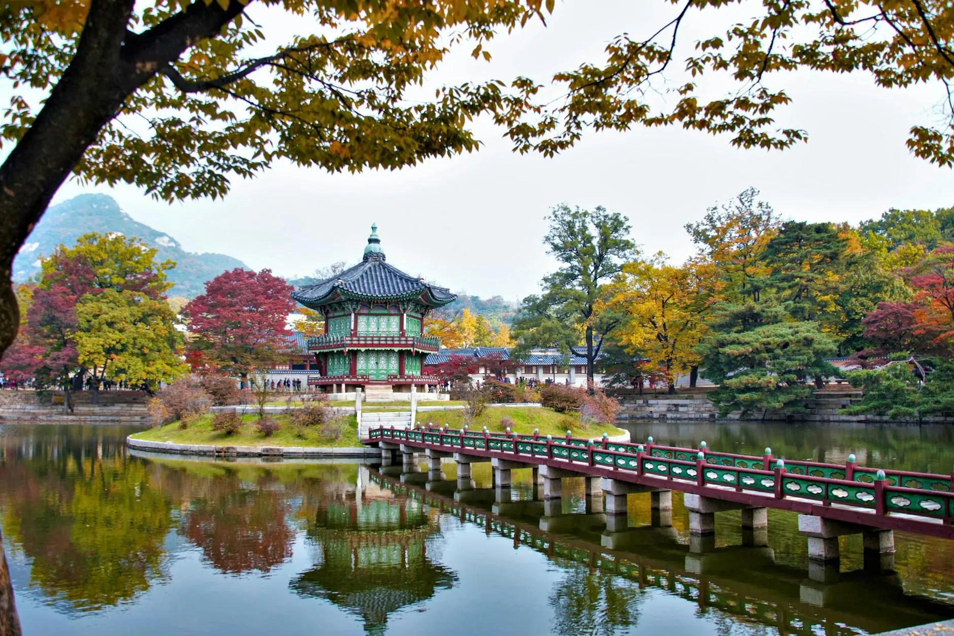 A peaceful view of Gyeongbokgung Palace surrounded by colorful autumn foliage, featuring a traditional pavilion and a serene pond reflecting the landscape.