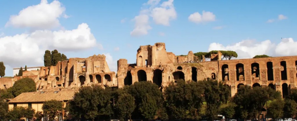 Panoramic view of the ancient Roman ruins in Rome, featuring weathered stone structures and trees against a blue sky with clouds.