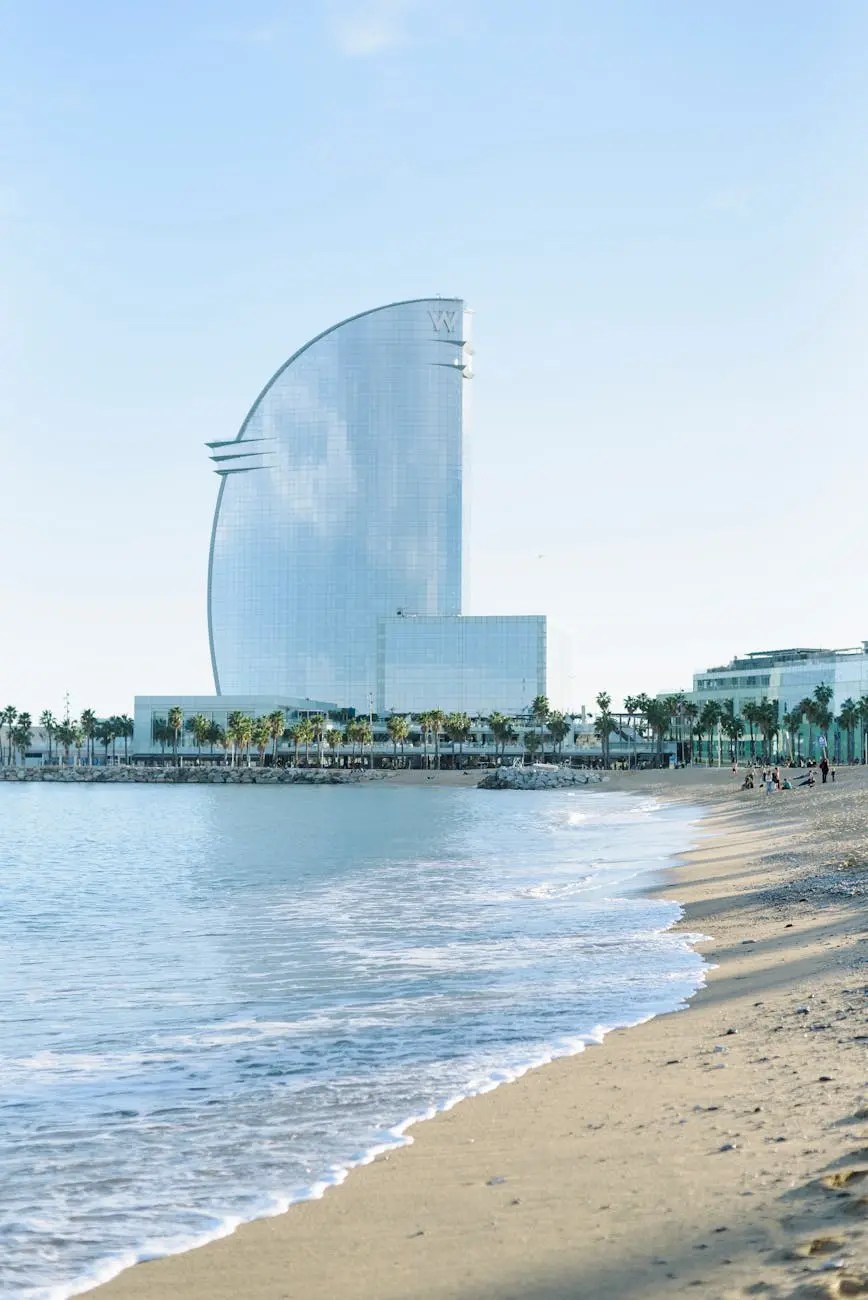 View of the W Barcelona hotel from the beach, featuring its distinctive sail-like shape and the shoreline with gentle waves, palm trees, and a clear sky.