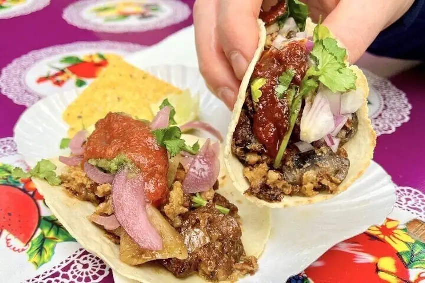 Two tacos filled with savory meat, topped with onions, cilantro, and sauces, held by a hand, with colorful table decor in the background.
