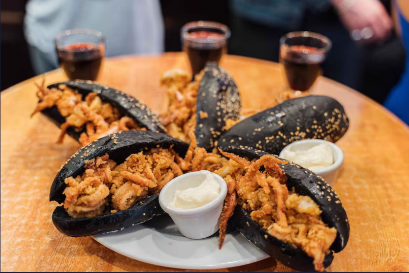 A plate of black sesame buns filled with fried seafood, accompanied by two small bowls of sauce and several glasses of red wine in the background.