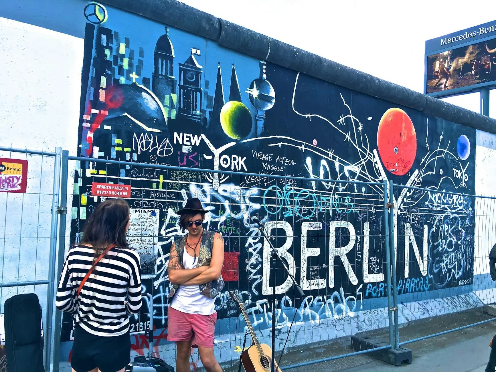 Two individuals interacting in front of a colorful mural on a section of the Berlin Wall, featuring vibrant graffiti and the word 'BERLIN'.
