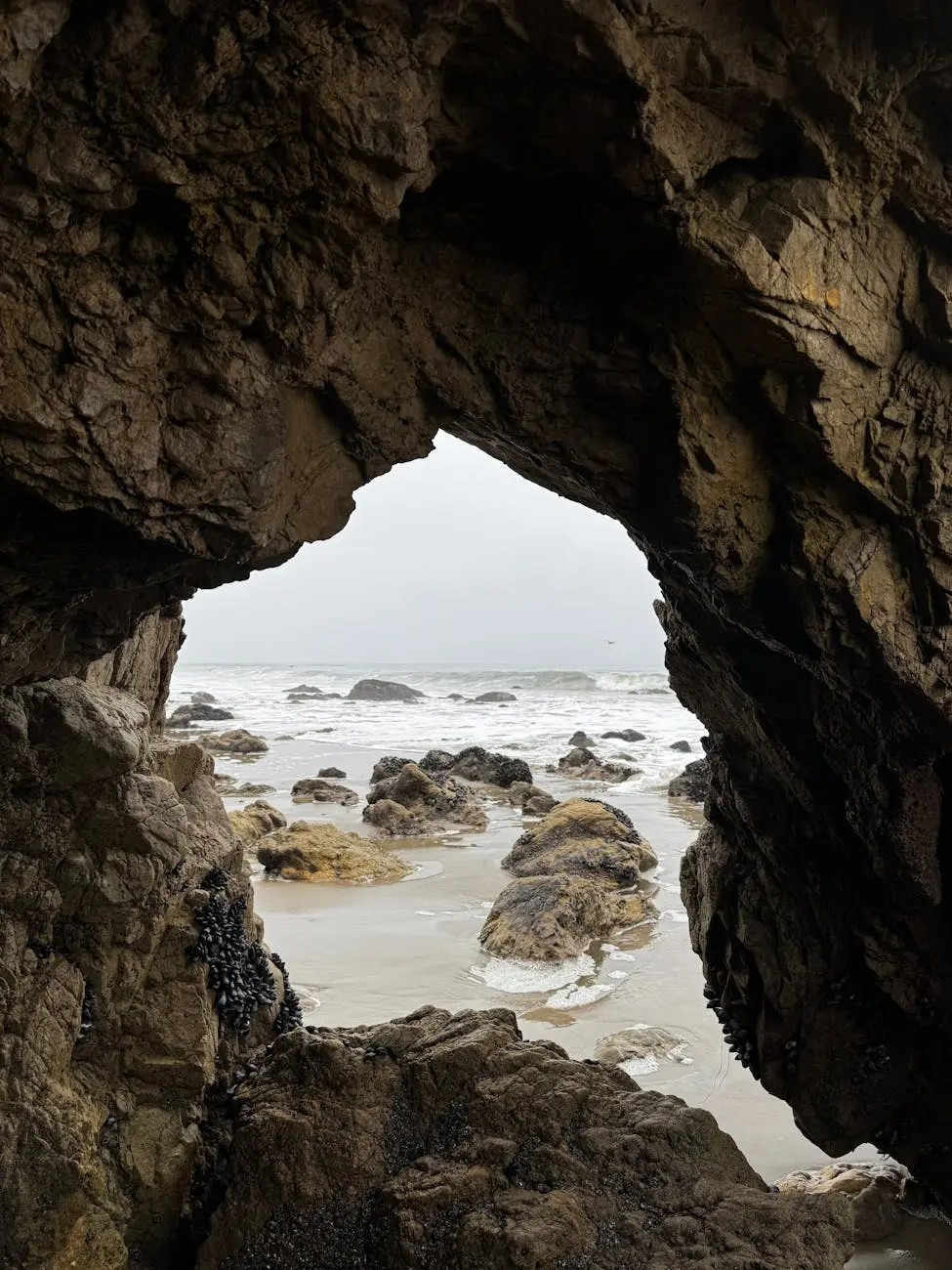 Vista de un agujero en una roca que da al mar, con olas y rocas en la playa al fondo.
