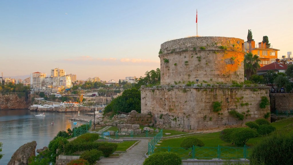 A historic stone tower with a Turkish flag on top, surrounded by lush greenery and views of the Mediterranean Sea in Antalya during sunset.