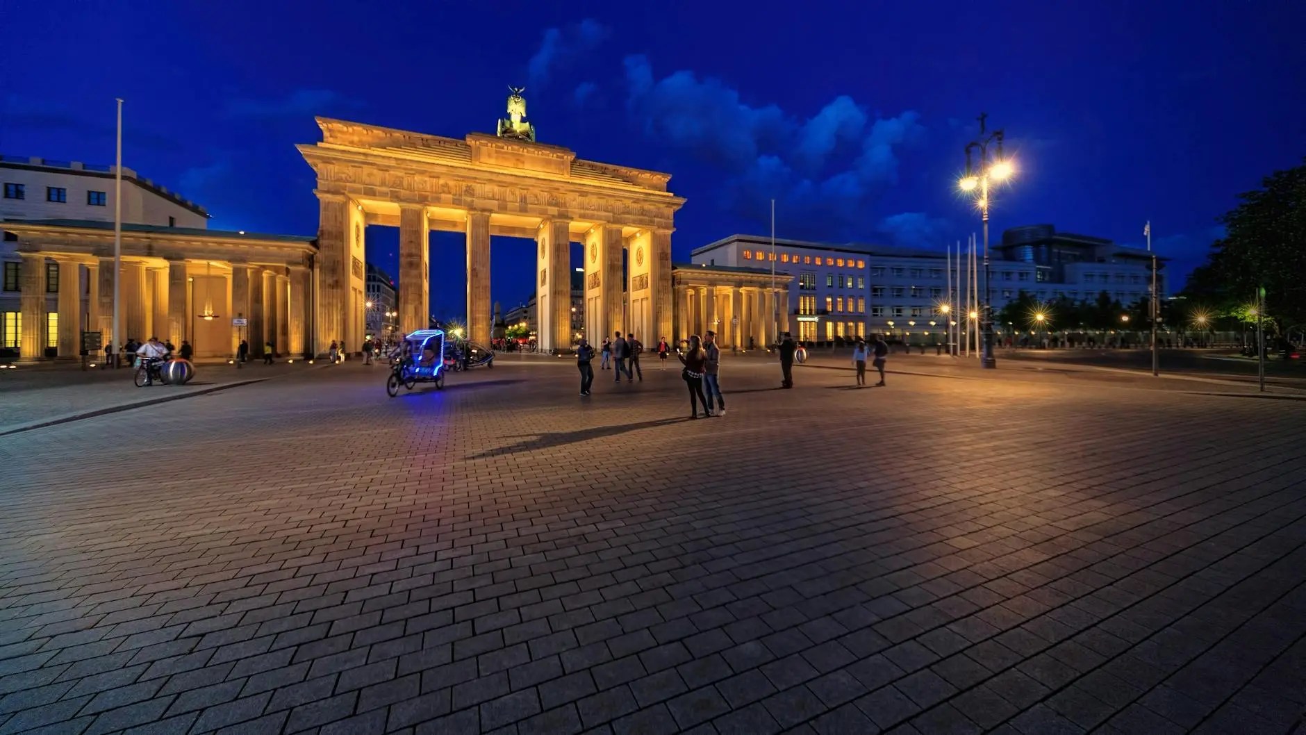 The Brandenburg Gate illuminated at night, surrounded by people and a clear blue sky.