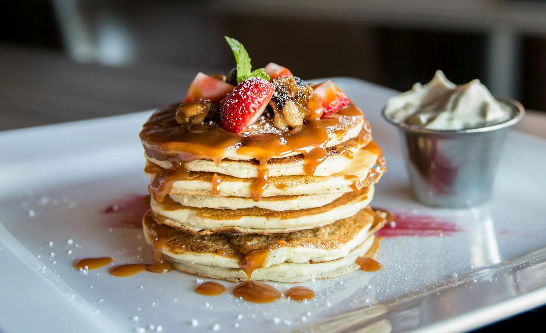 A stack of pancakes topped with sliced strawberries, caramel drizzle, and a sprinkle of powdered sugar, accompanied by a small cup of whipped cream.