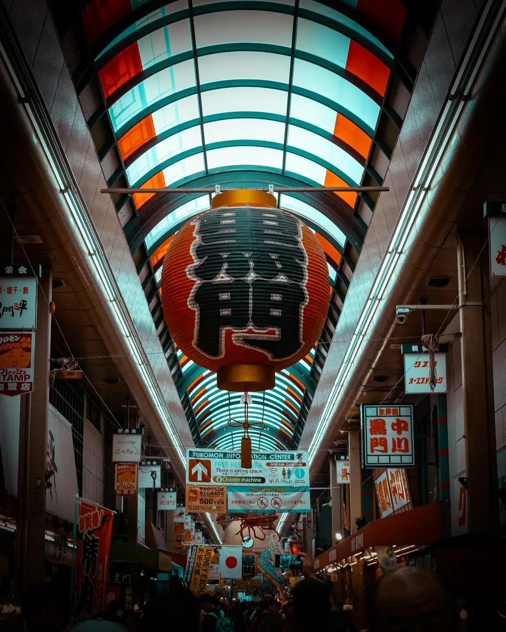 A bustling scene inside Kuromon Ichiba Market in Osaka, featuring overhead colorful banners and large lanterns, with a crowd of shoppers enjoying the vibrant atmosphere.