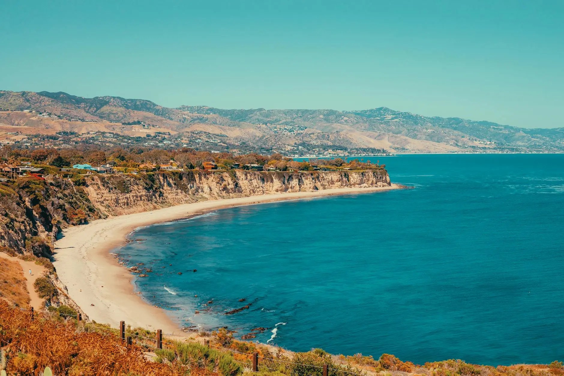 A scenic view of a coastal cliff with sandy beach and turquoise water under a clear blue sky, showcasing the natural beauty of the Los Angeles coastline.