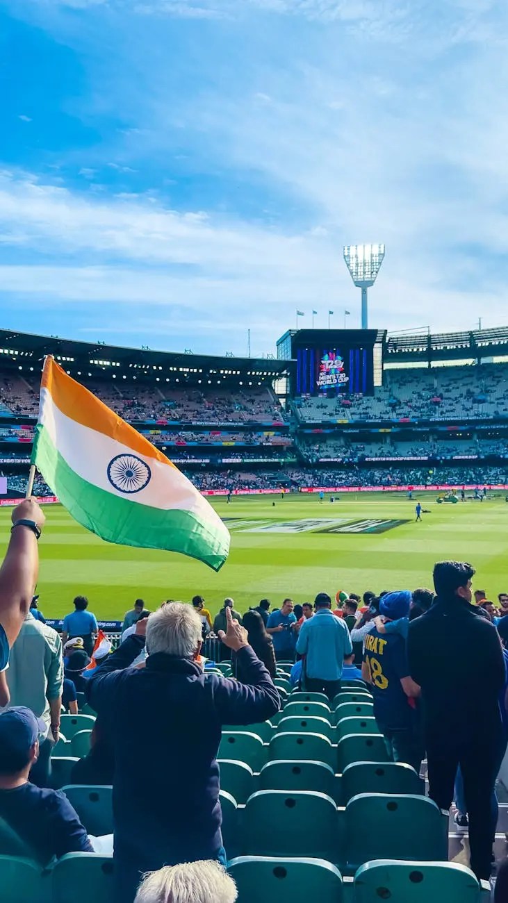 Crowd at the Melbourne Cricket Ground waving an Indian flag during a cricket match, with seats and players visible in the background.