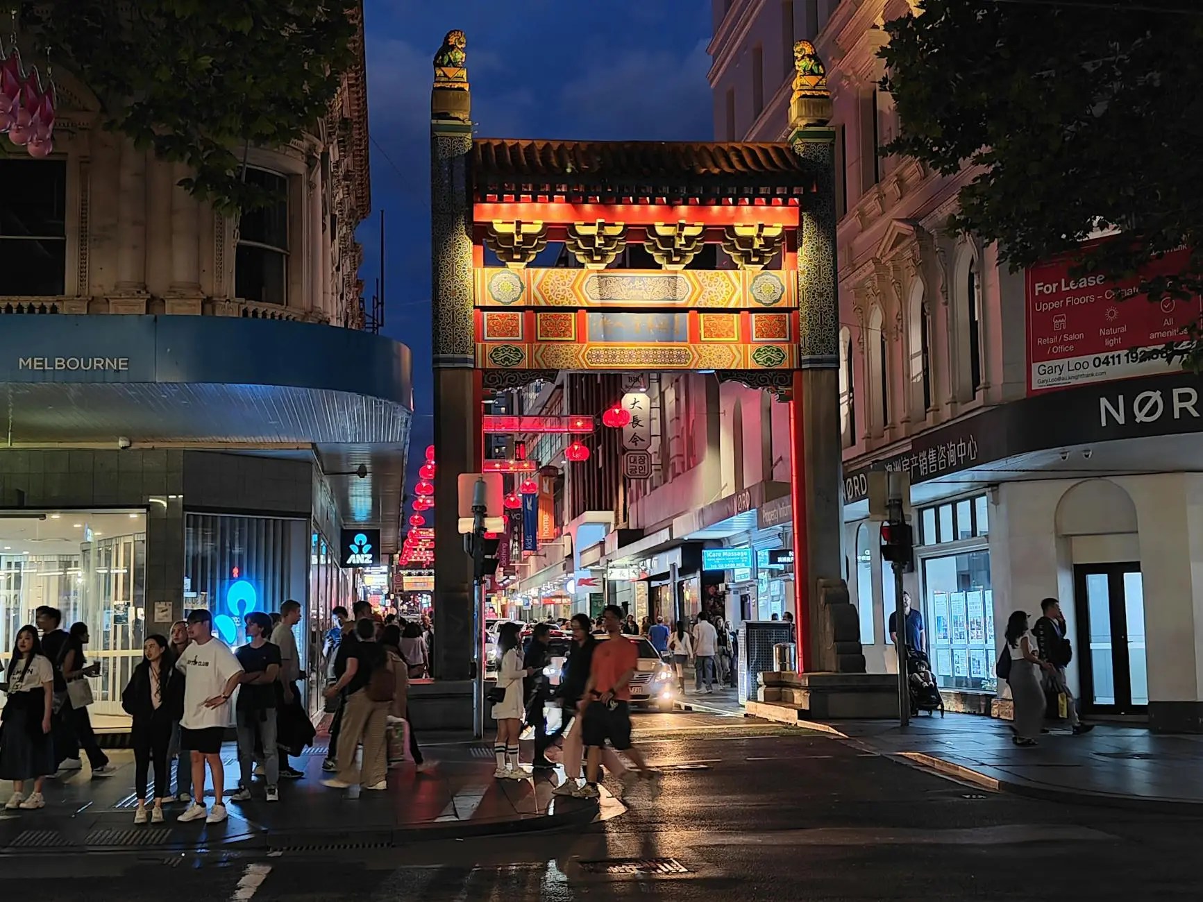 A bustling street scene in Melbourne's Chinatown at night, featuring a vibrant archway adorned with traditional Chinese decorations and lanterns, surrounded by pedestrians enjoying the lively atmosphere.