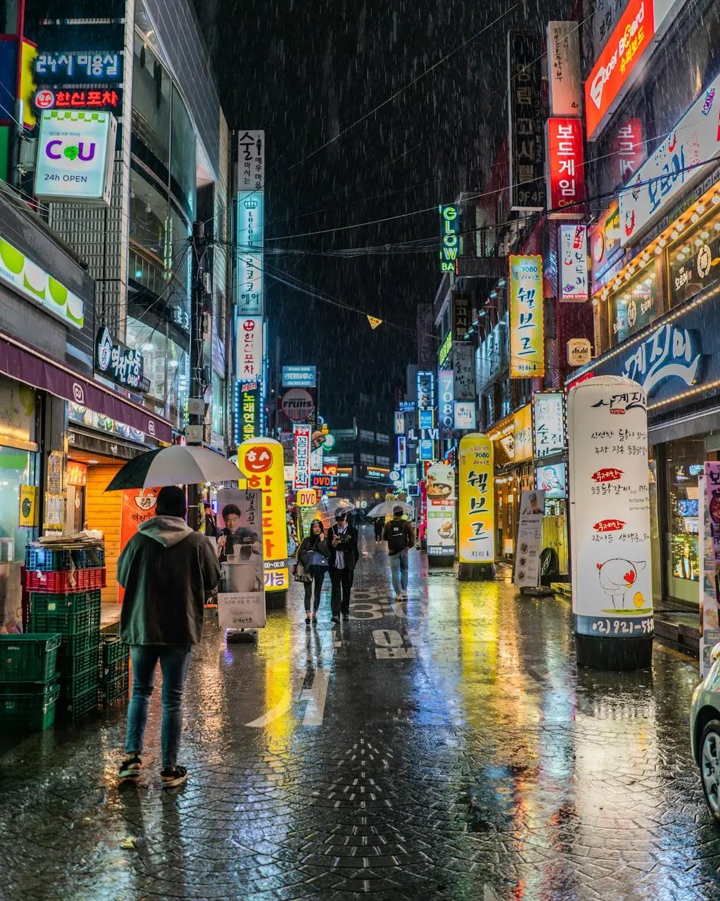 A bustling street in Seoul at night, illuminated by vibrant neon signs and reflections on wet pavement, with people walking under umbrellas in a light rain.
