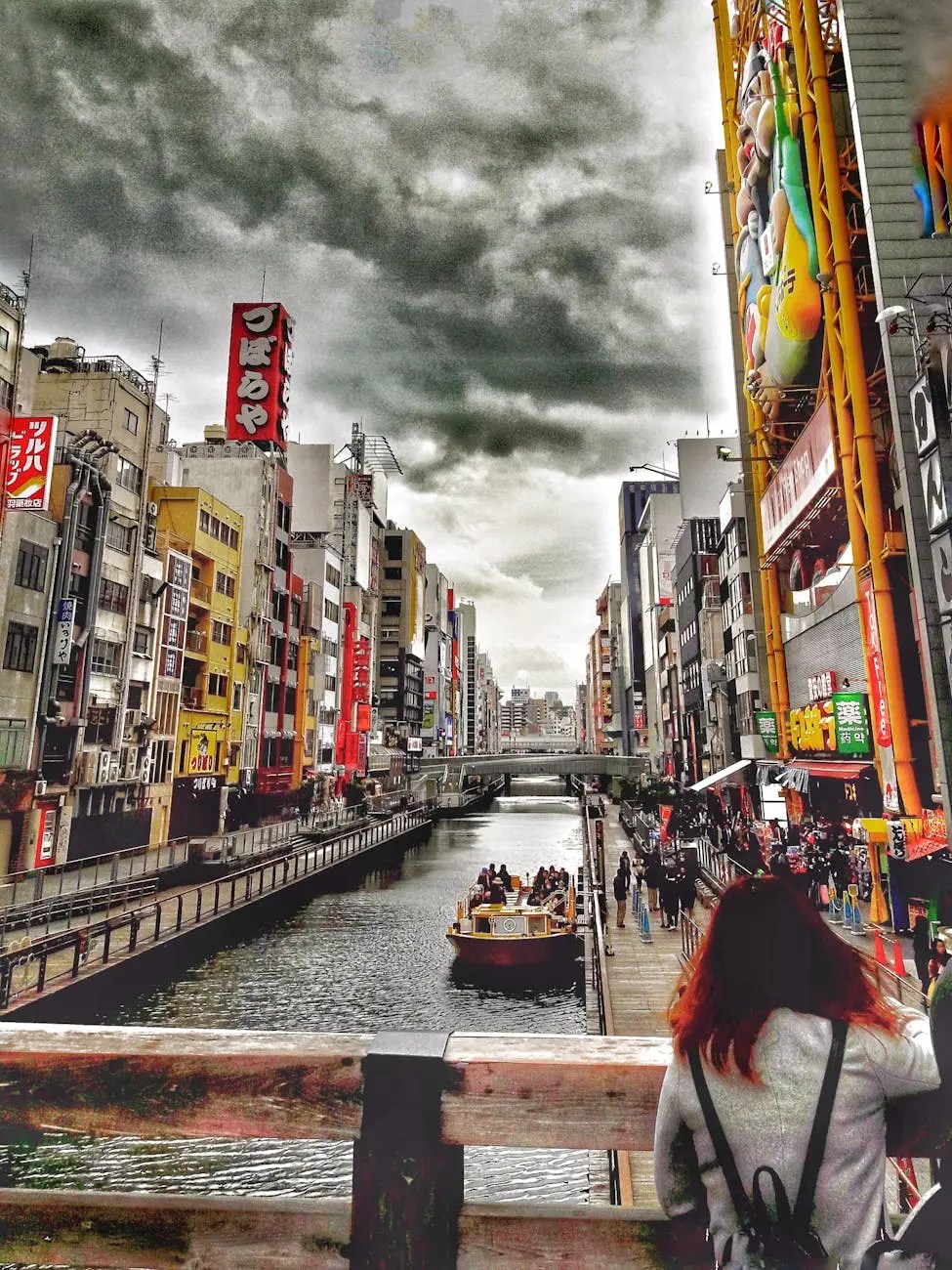 A person observes the Dotonbori area, featuring a bustling canal with a sightseeing boat and vibrant buildings adorned with colorful advertisements and neon signs under a cloudy sky.