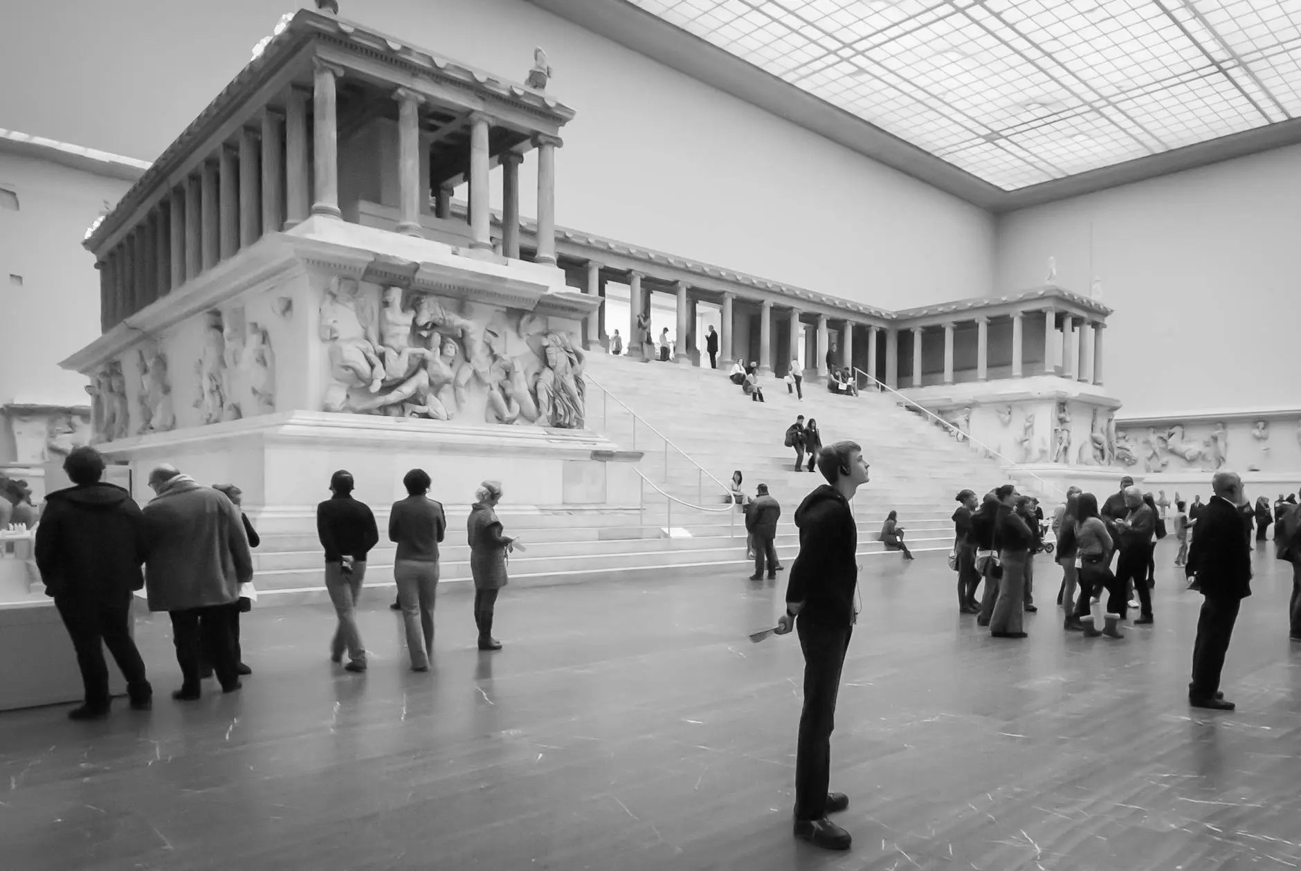 A black and white image of visitors inside a large museum, featuring an ancient structure resembling a temple on a raised platform, with multiple staircases and detailed sculptures. People are scattered throughout the space, some looking at the artwork and others standing in contemplation.