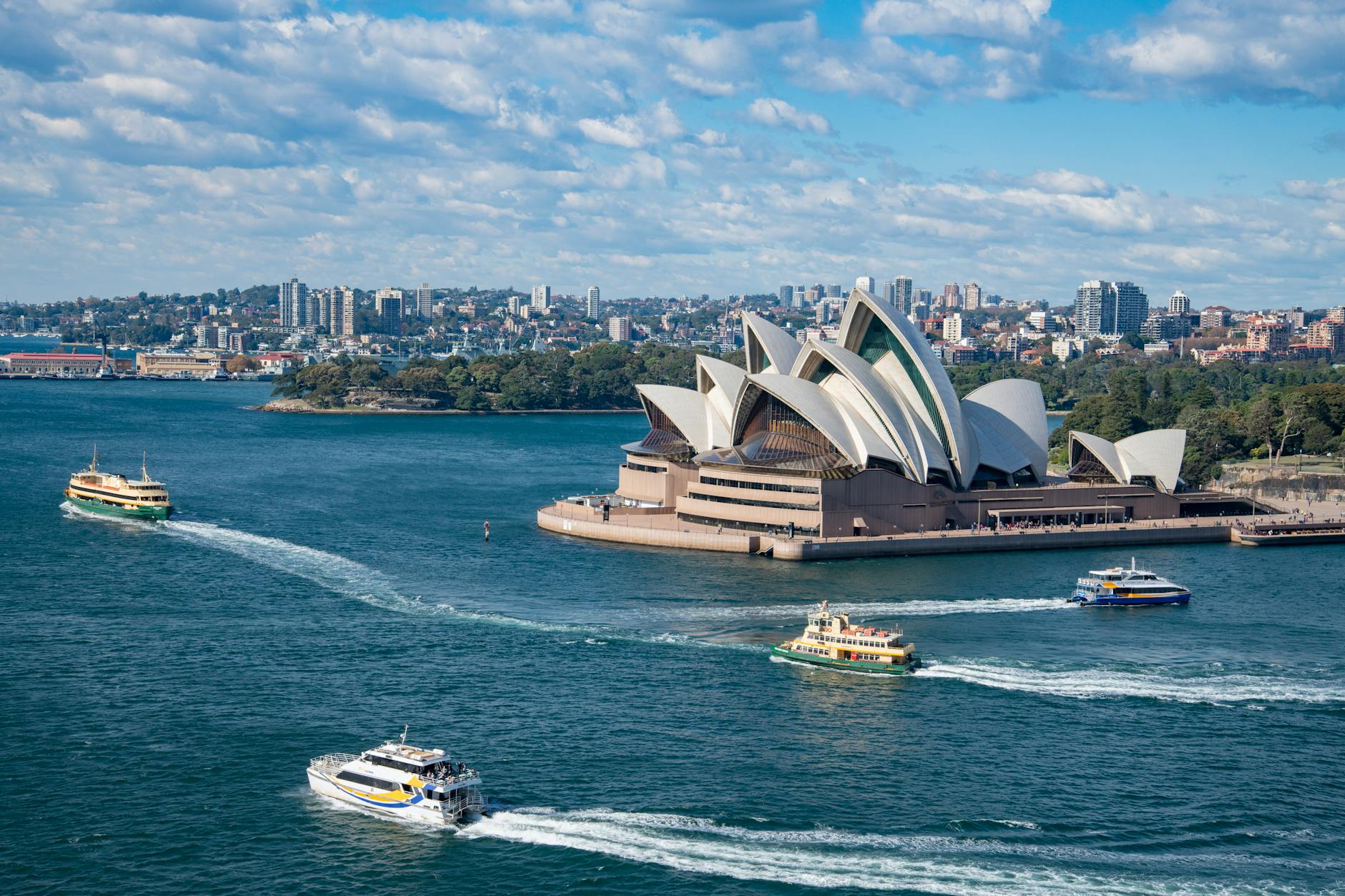 Aerial view of the Sydney Opera House surrounded by water, with several ferries traversing the harbor and the Sydney skyline in the background.
