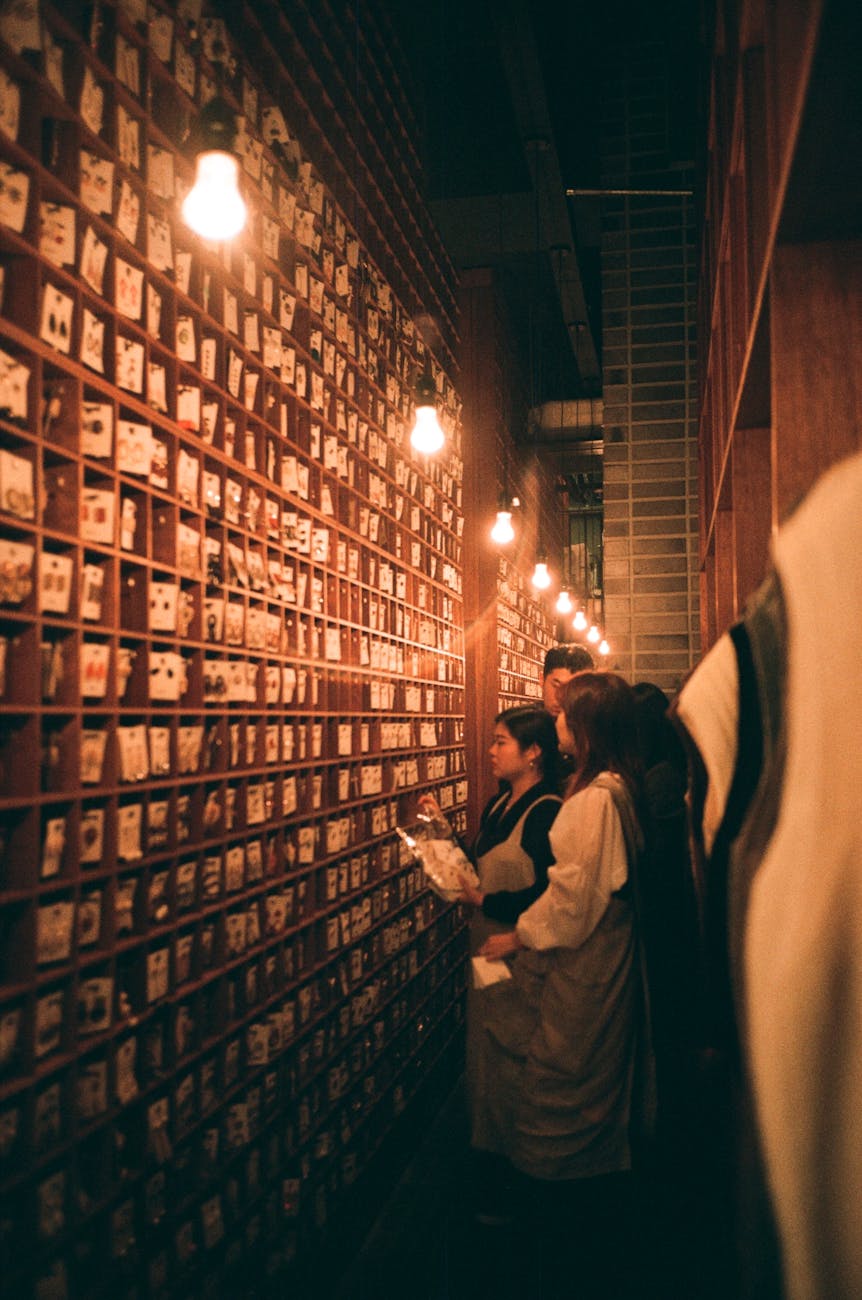 A group of people examining various items displayed on a wooden wall covered in small compartments, illuminated by warm overhead lights.