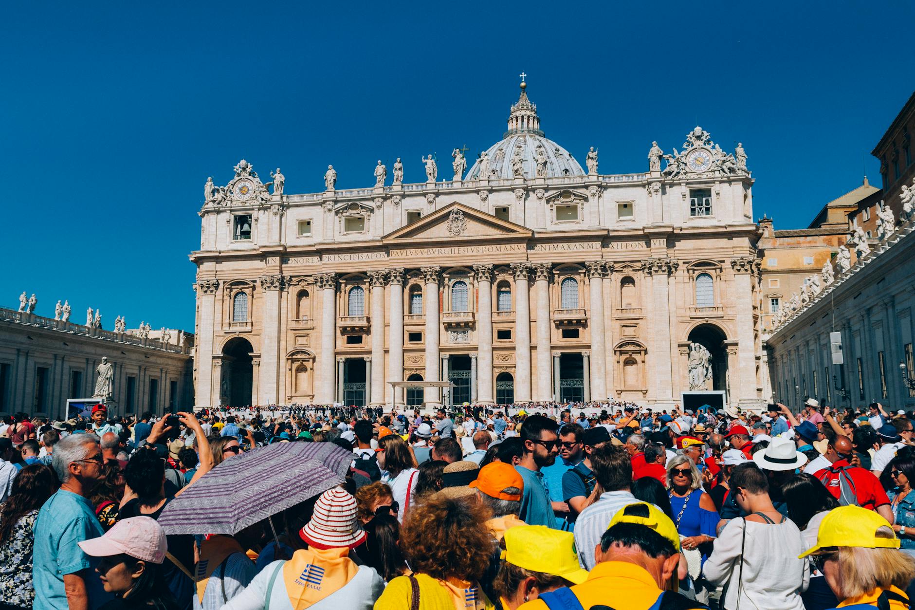 Crowd of tourists gathered in front of St. Peter's Basilica in Vatican City, with the magnificent façade and dome visible against a clear blue sky.