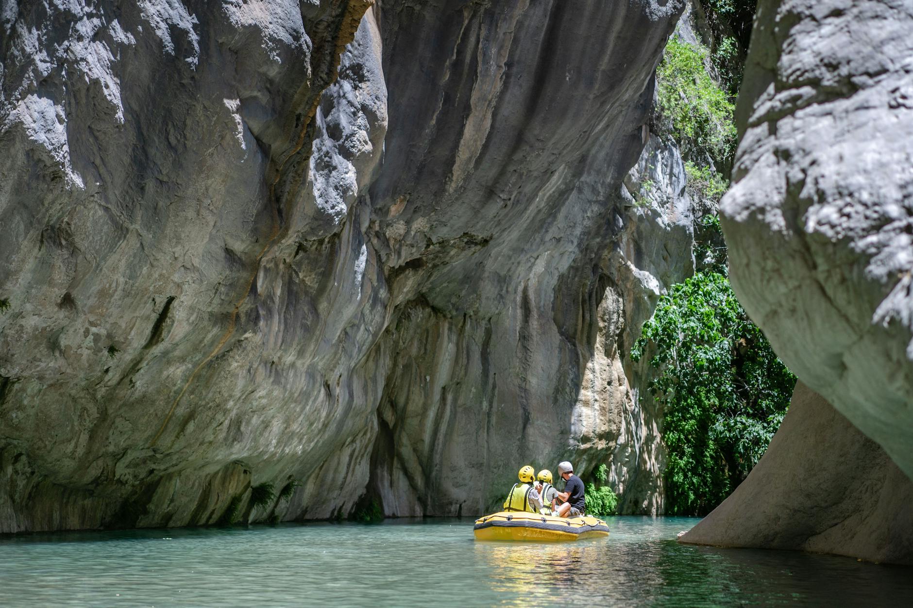 A group of four people in a yellow inflatable raft navigating through a narrow canyon with steep, rocky walls and greenery surrounding the water.