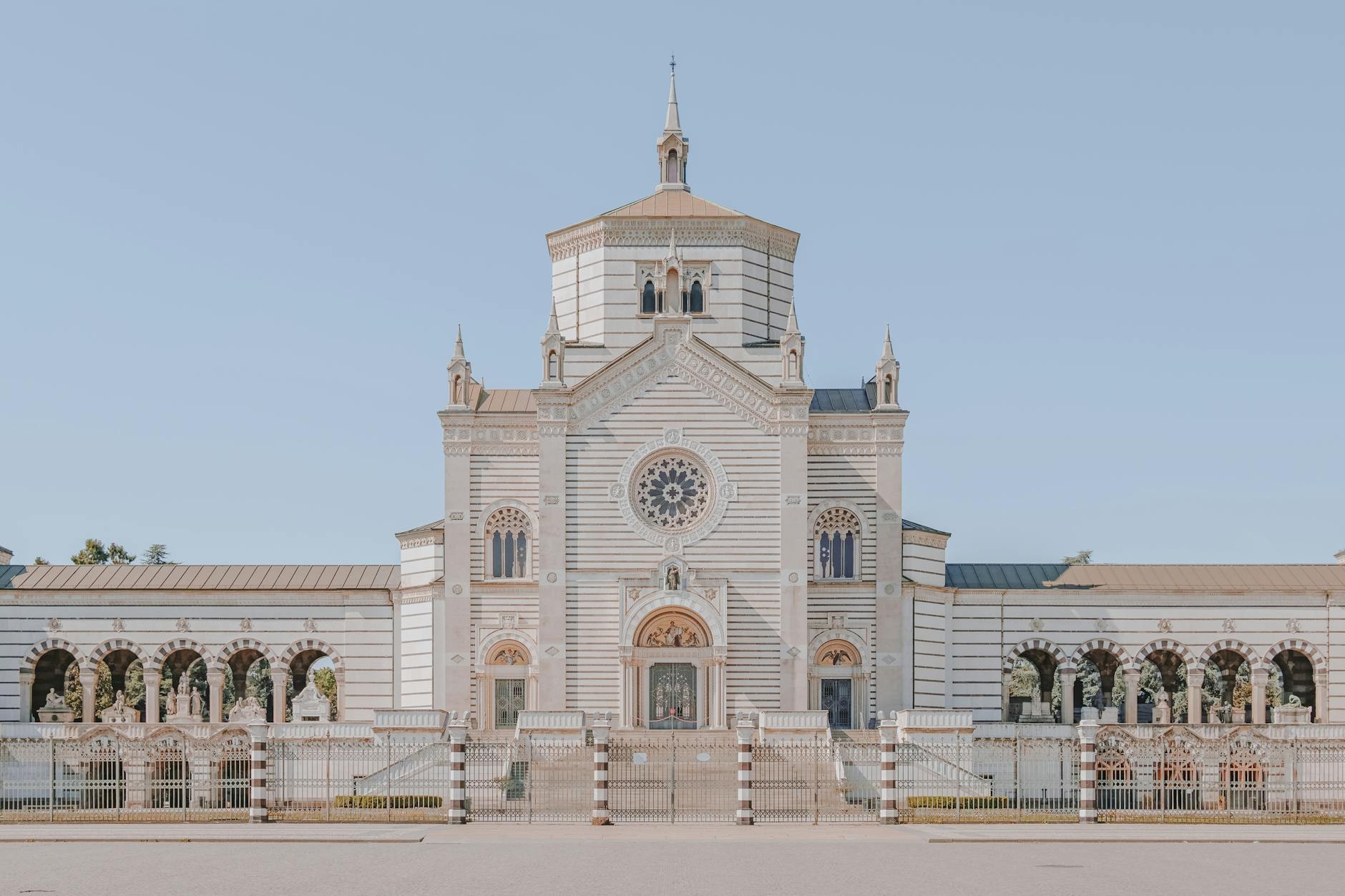 Facade of the Cimitero Monumentale in Milan, showcasing its ornate architecture and decorative elements against a clear blue sky.