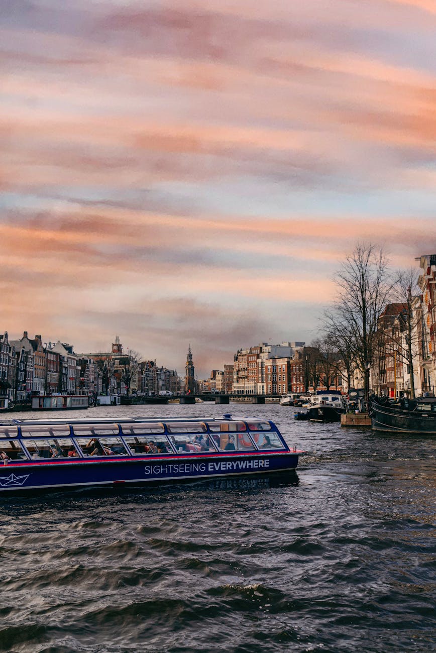 A sightseeing boat navigates through Amsterdam's canals during sunset, showcasing picturesque buildings and trees along the waterfront.