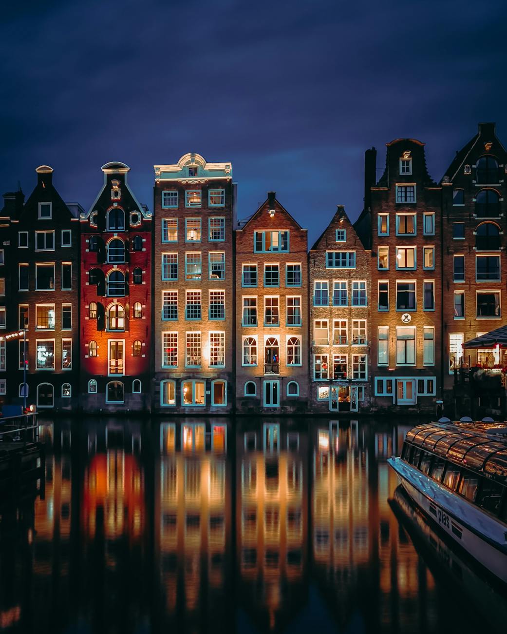 A scenic view of illuminated historic buildings along a canal in Amsterdam at night, with reflections in the water.