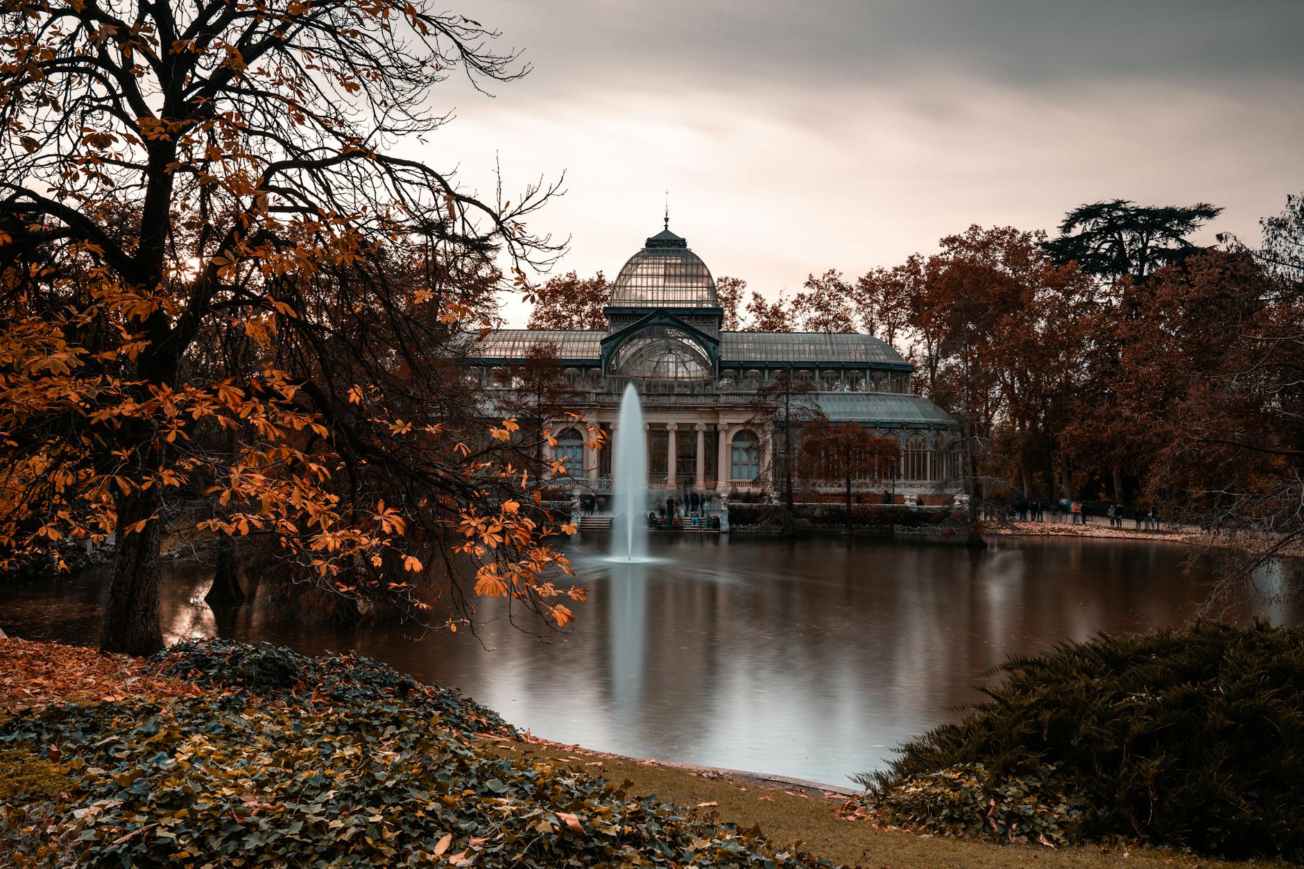 View of the Crystal Palace in Retiro Park, Madrid, surrounded by autumn leaves and a serene pond with a fountain.