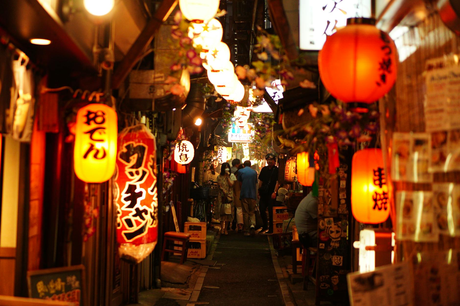 A narrow alley in Tokyo filled with vibrant lanterns and signage, creating a cozy, inviting atmosphere for diners. People can be seen walking and enjoying the lively atmosphere of this bustling street.