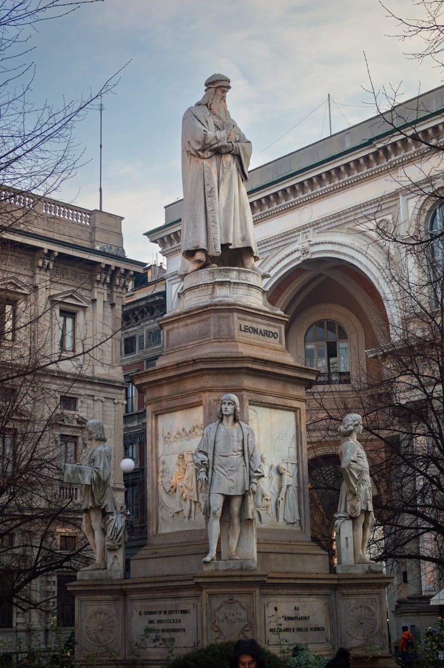 Statue of Leonardo da Vinci surrounded by additional sculptures, located in a square in Milan, Italy.