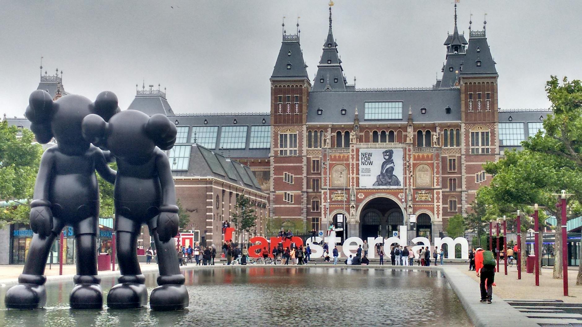 Outdoor view of the Rijksmuseum in Amsterdam with large black sculptures of two figures in front and tourists nearby.