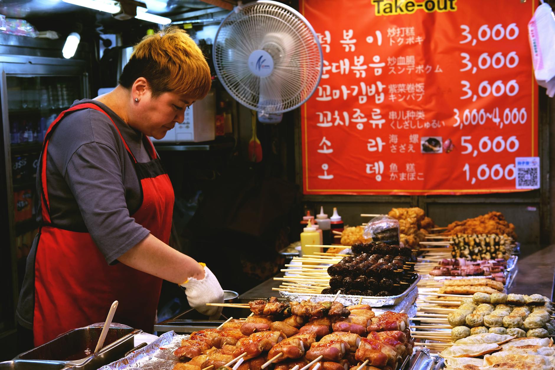 A vendor preparing skewered street food at a market in Seoul, with various food items displayed on a counter and a bright take-out menu in the background.