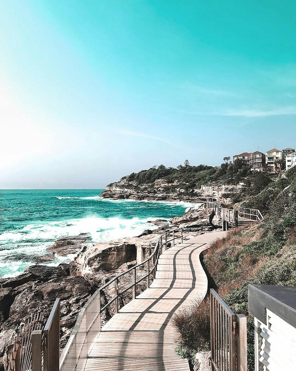 A scenic coastal walkway with wooden paths along rocky cliffs and turquoise ocean waves, leading toward seaside homes in Sydney, Australia.