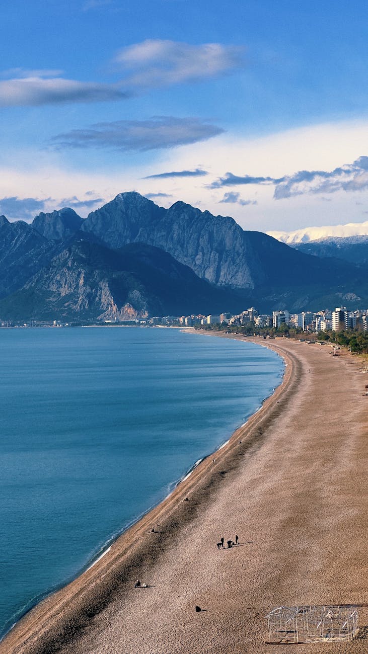 Konyaaltı Beach – Where Golden Sands Meet the Turquoise Mediterranean" A panoramic view of Konyaaltı Beach in Antalya, Turkey, featuring golden sandy shores, turquoise Mediterranean waters, and the Taurus Mountains in the background. A few people can be seen walking along the beach.