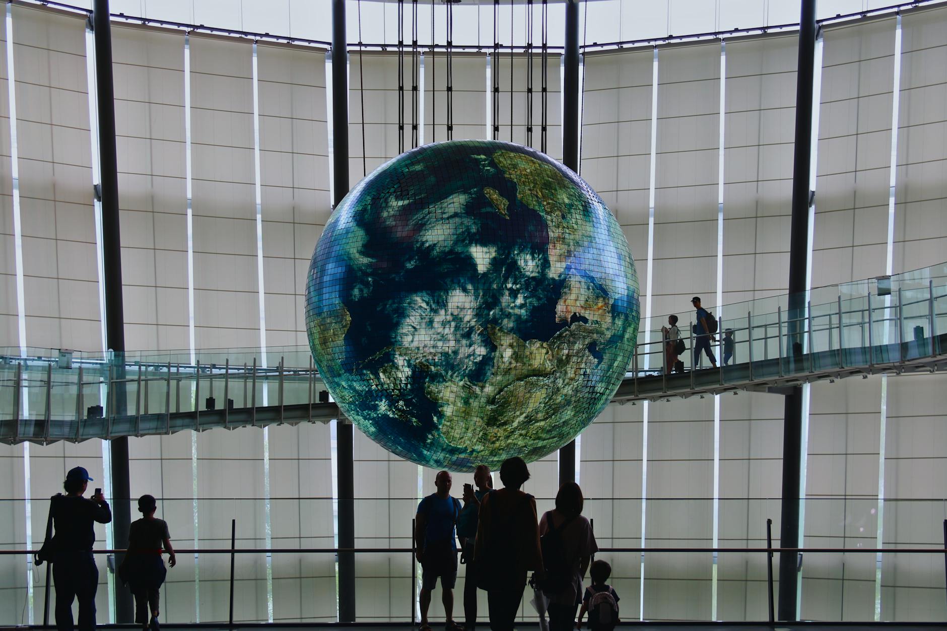 A large illuminated globe hanging in a modern museum, with silhouettes of visitors walking along a bridge in the background.
