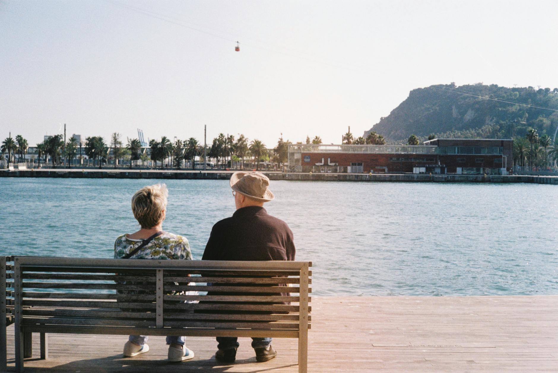 A couple sitting on a wooden bench by the water in Barcelona, looking out at the sea and nearby palm trees.
