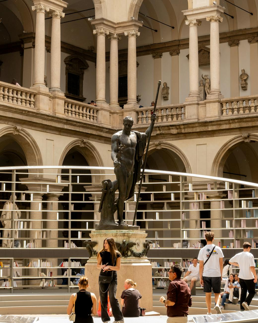 Statue of a historical figure in a spacious courtyard with columns, surrounded by visitors. People are sitting and standing in various poses, enjoying the ambiance.