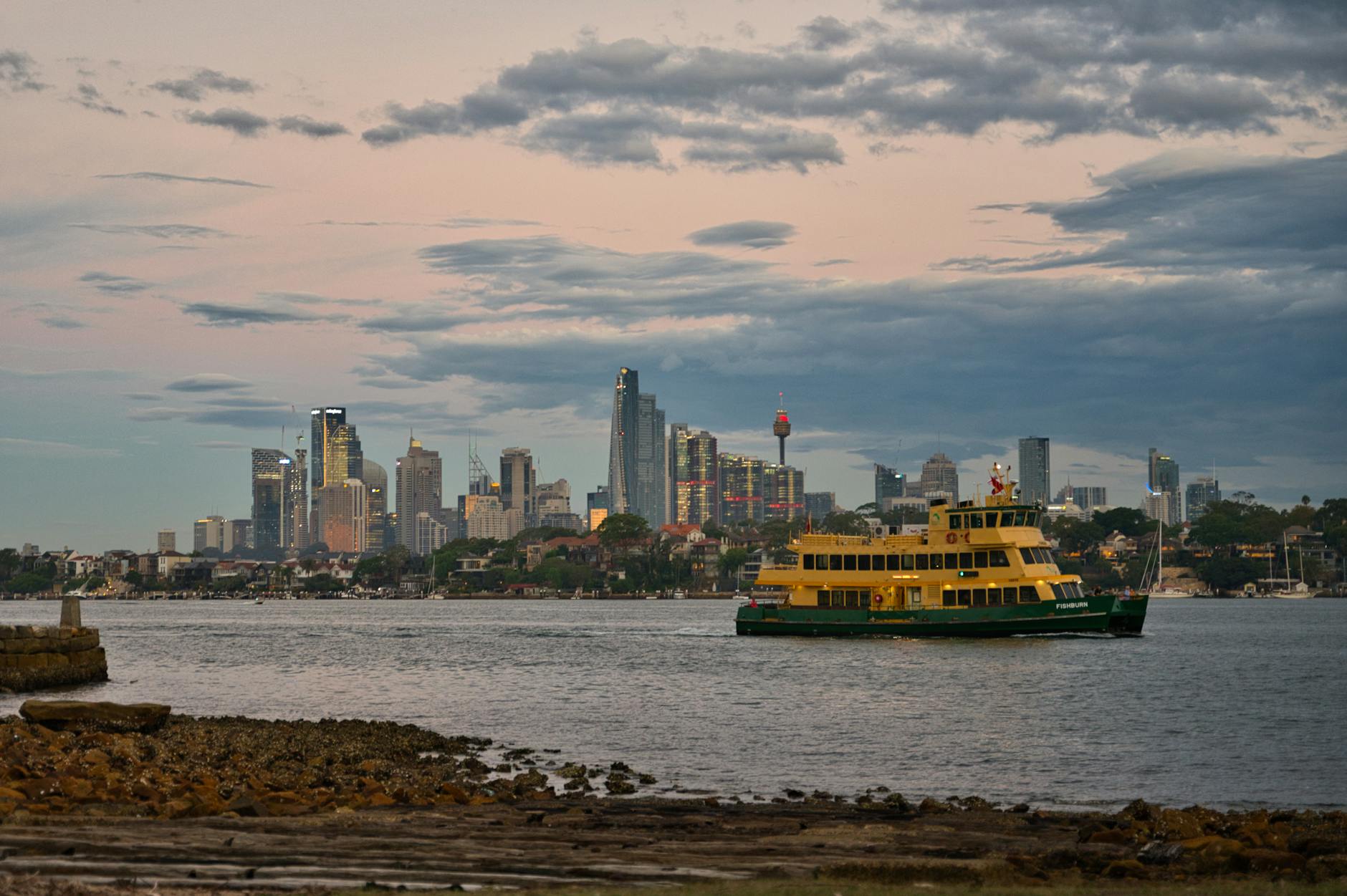 A ferry cruising on the water with the Sydney skyline in the background, featuring tall buildings under a cloudy sky during sunset.
