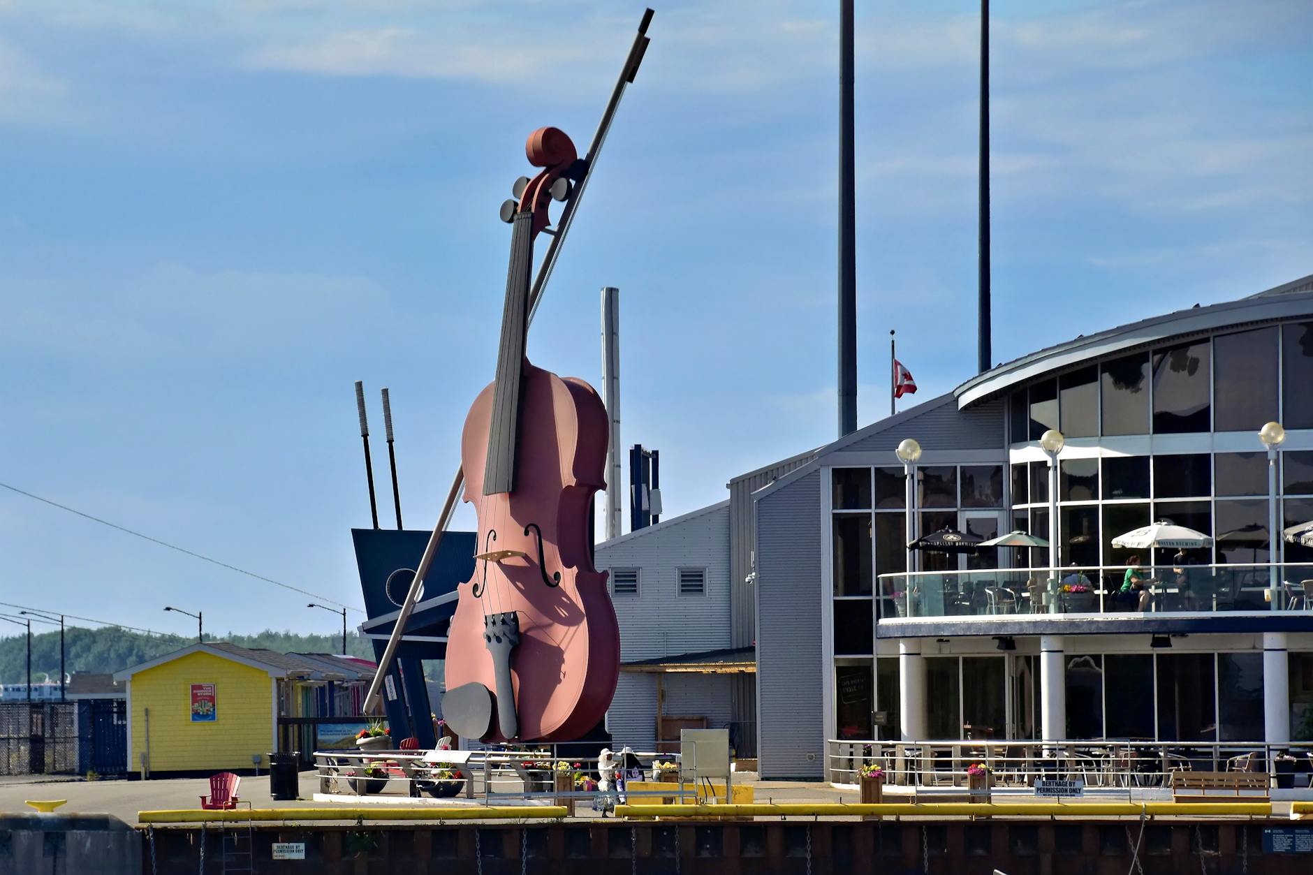A large, pink violin sculpture positioned near a modern building with a terrace, set against a clear blue sky.