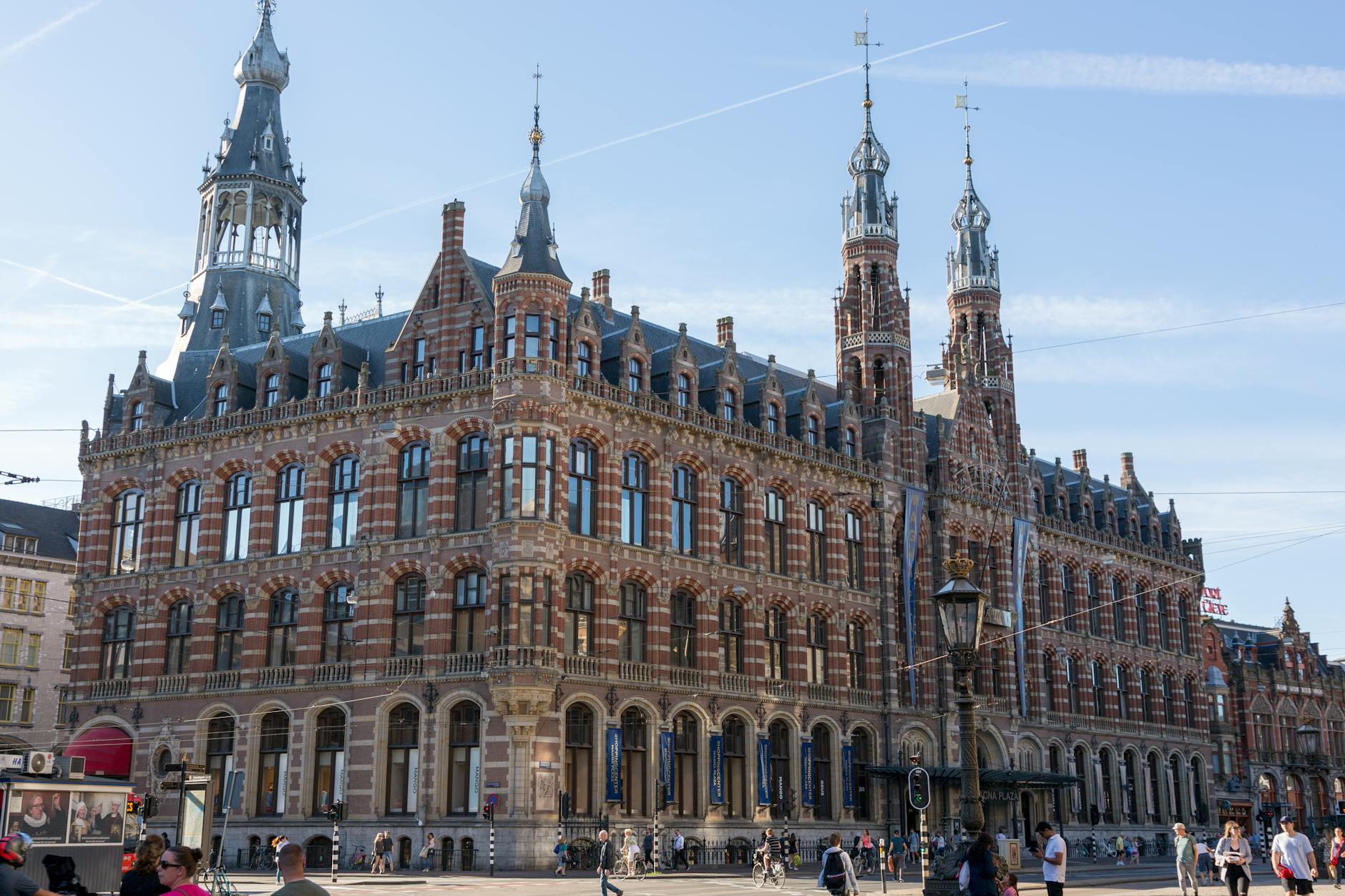 A detailed view of a historic building in Amsterdam featuring ornate architecture and tall spires, with passersby and bicycles in the foreground.