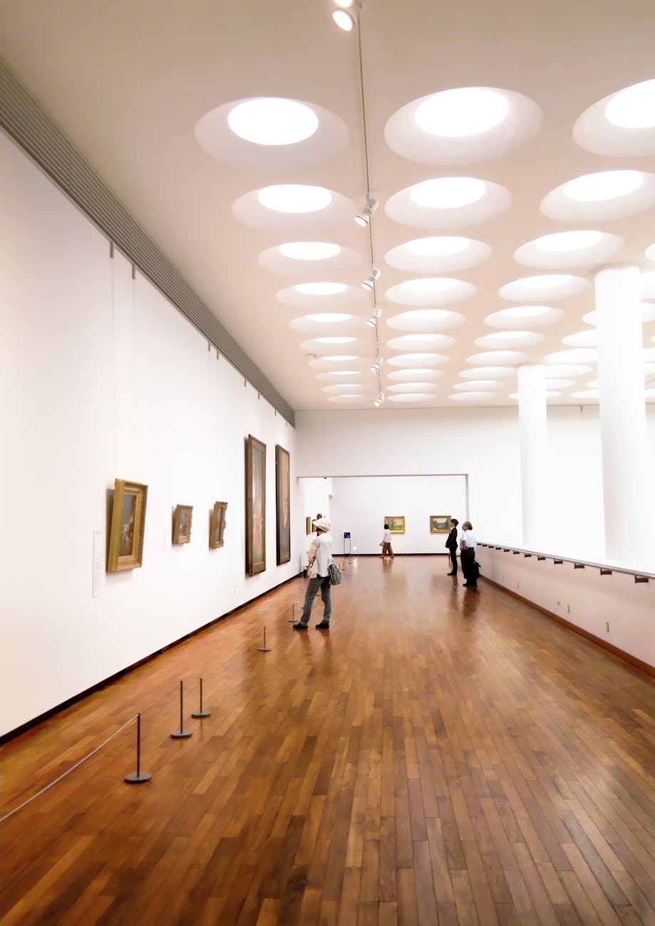 Interior view of an art gallery featuring wooden flooring, modern lighting, and several paintings displayed on white walls. Visitors are observing the artwork.