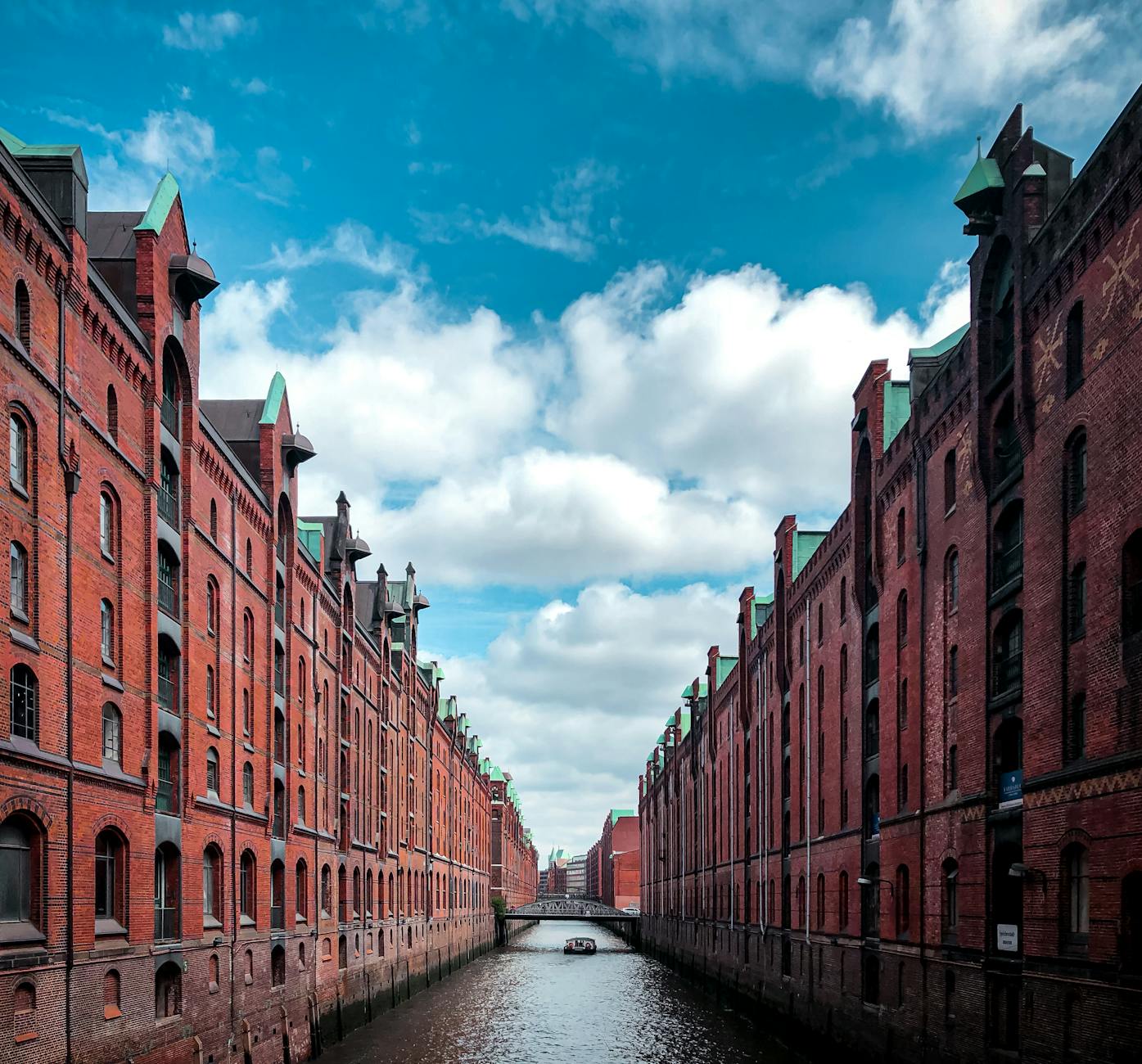 A scenic view of a canal flanked by historic red brick warehouses under a blue sky with scattered clouds.