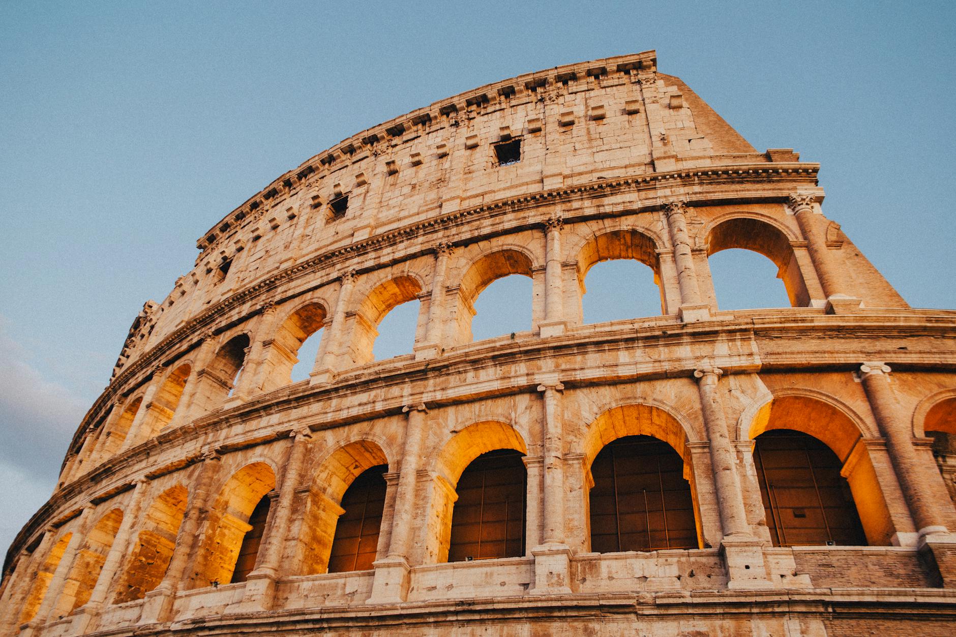 A low-angle view of the Colosseum in Rome, highlighting its ancient arches and illuminated stonework against a clear blue sky.