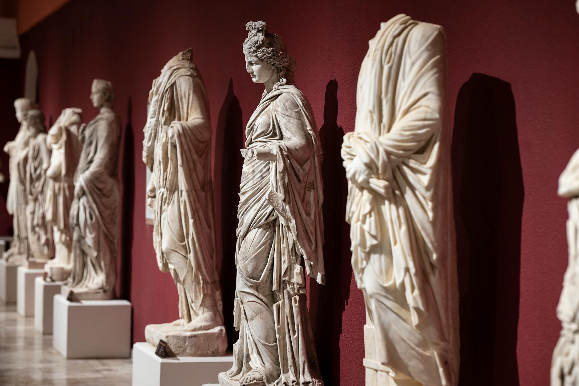 "Whispers of Antiquity – Timeless Statues at Antalya Museum" A row of ancient marble statues in an exhibition, showcasing the fine details of their draped clothing and features, set against a deep red wall.